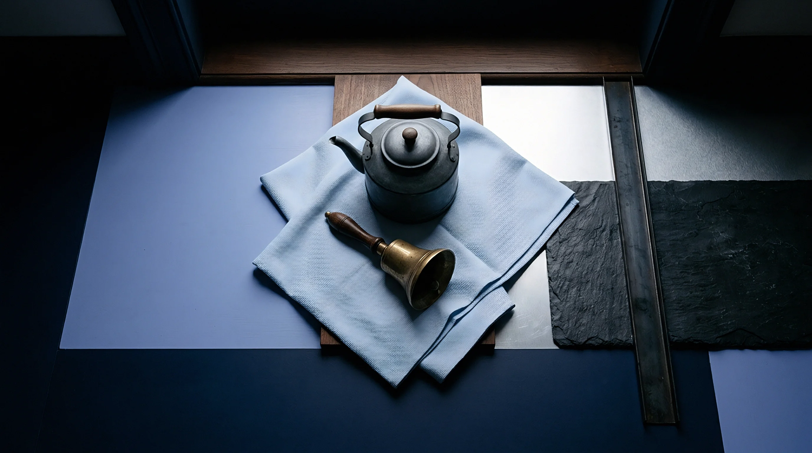 An aged metal kettle and a brass bell rest on light blue textile over varied raw material samples and industrial surfaces.