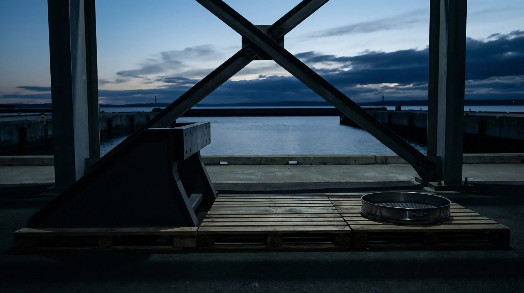 Heavy steel structural supports frame wooden pallets holding industrial metal components on a concrete maritime loading dock overlooking harbor waters.